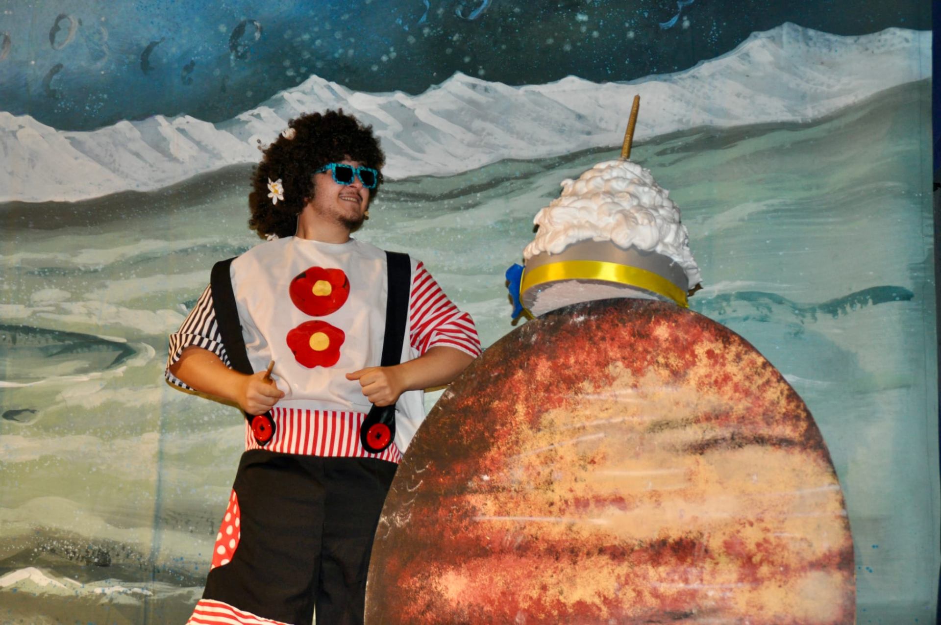 Personaggio con parrucca afro e occhiali davanti a pianeta di scena